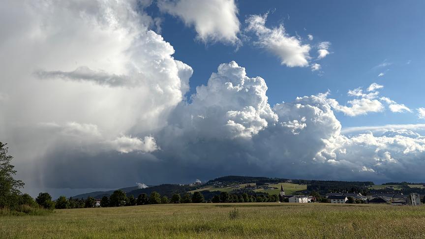 Ein weitläufiges Landschaftsfoto unter einem dramatischen Sommerhimmel. Die obere Bildhälfte wird von massiven, kontrastreichen Wolkenformationen dominiert. Links baut sich eine riesige, weiße Cumulonimbus-Wolke auf, aus deren dunkler Unterseite links außen ein sichtbarer Regenschauer auf die Erde fällt. Rechts davon türmen sich weitere, vom Sonnenlicht hell erleuchtete Quellwolken vor einem tiefblauen Himmel auf. Im unteren Drittel des Bildes erstreckt sich eine flache, grüne Wiese. Dahinter verläuft eine Hügelkette mit vereinzelten Bäumen und einer kleinen Ortschaft, in der ein spitzer Kirchturm aufragt.