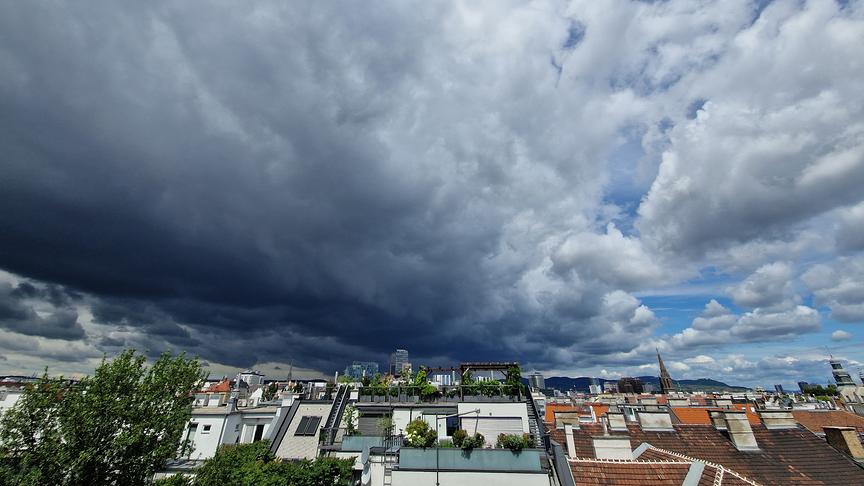 Eine dichte Decke aus kontrastreichen Wolken zieht über eine städtische Dachlandschaft. Im linken und unteren Bereich ballen sich bedrohliche, dunkelgraue Regenwolken zusammen. Zur rechten Seite hin reißt die Wolkendecke auf und gibt den Blick auf weißen, fluffigen Schönwetterwolken vor einem blauen Himmel frei. Am unteren Bildrand erstrecken sich die Dächer von Wohnhäusern mit Terrassen, vereinzelten Bäumen und Kirchtürmen in der Ferne.