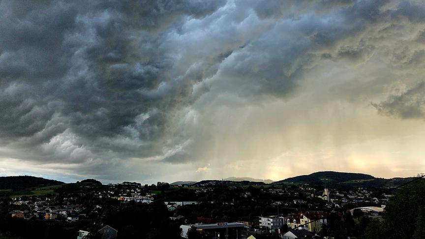 Eine weitwinklige Landschaftsaufnahme zeigt eine dramatische, stürmische Wolkenformation über einer Kleinstadt und hügeliger Landschaft bei Tageslicht.