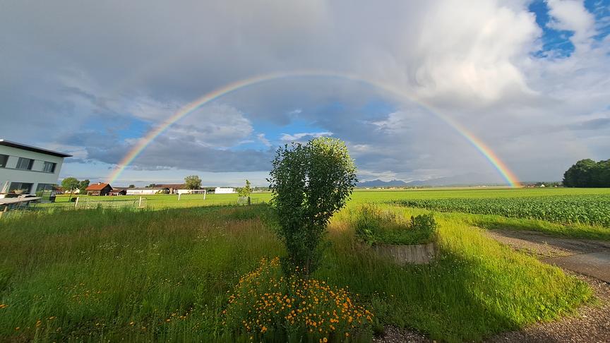 Eine weite Landschaftsaufnahme unter einem teilweise bewölkten Himmel mit einem großen, perfekt geformten Regenbogen.