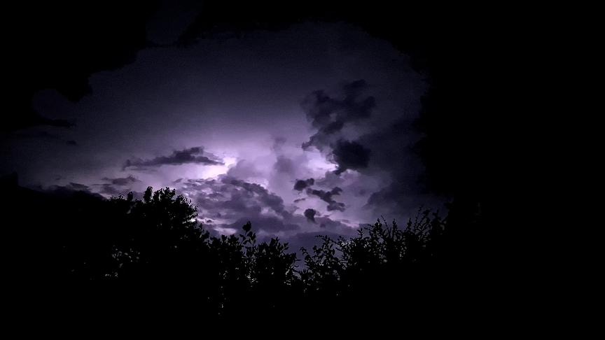 Helles Wetterleuchten erhellt Wolken in der Dunkelheit. Im Vordergrund sind einige Silhouetten von Bäumen zu sehen.