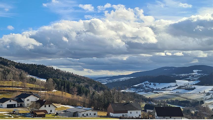Hügel und Tal mit Häusern sind teils noch leicht verschneit, drüber hohe Wolkenformationen