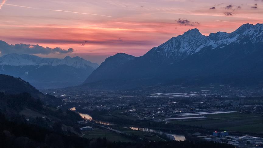 Ein Tal mit Fluss umgeben von hohen Bergformationen wird von der hellen Dämmerung in ein rötliches Licht getaucht.