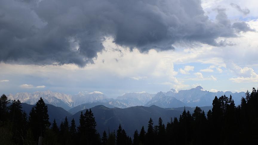 © Walter Hochfellner Vordergrund die Silhouette von den Baumkronen eines Waldes. Im Hintergrund erstreckt sich ein weitläufiges Bergpanorama. Die Szenerie wird von dunkles, großen Wolken bedeckt, die eine dunkle Stimmung verbreiten.