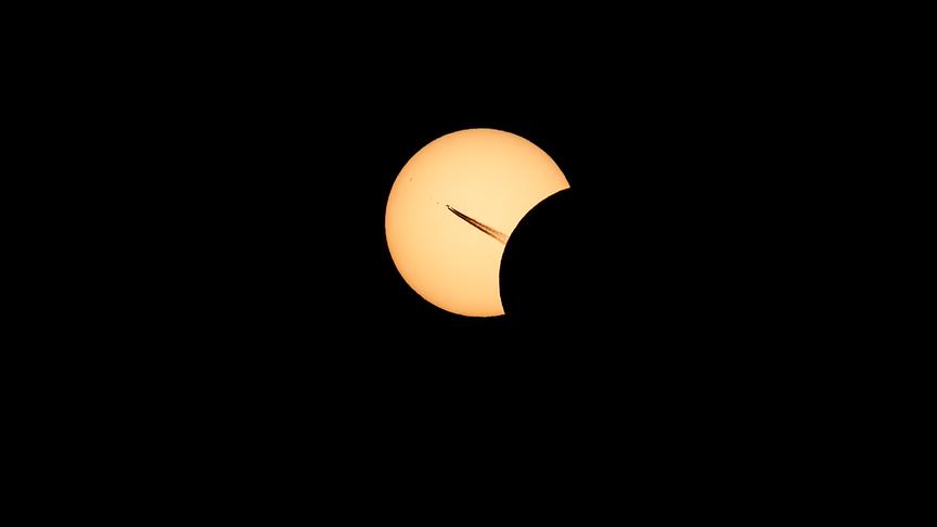 Nahaufnahme der partiellen Sonnenfinstenis. Der Mond schiebt sich dunkel vo die Sonne, während ein Flugzeug optisch perfekt vor der Sonne vorbeifliegt.