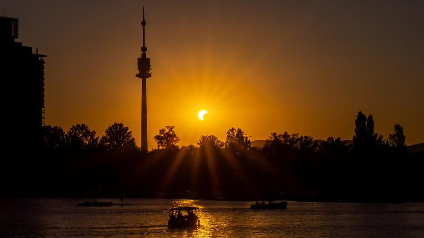 Die partielle Sonnenfinsternis taucht das Gewässer der alten Donau in warm-oranges Licht. Ein paar Leute sind dort mit Boot unterwegs. Im Hintergrund zeichnen sich Bäume und sehr präsent der Donauturm ab.