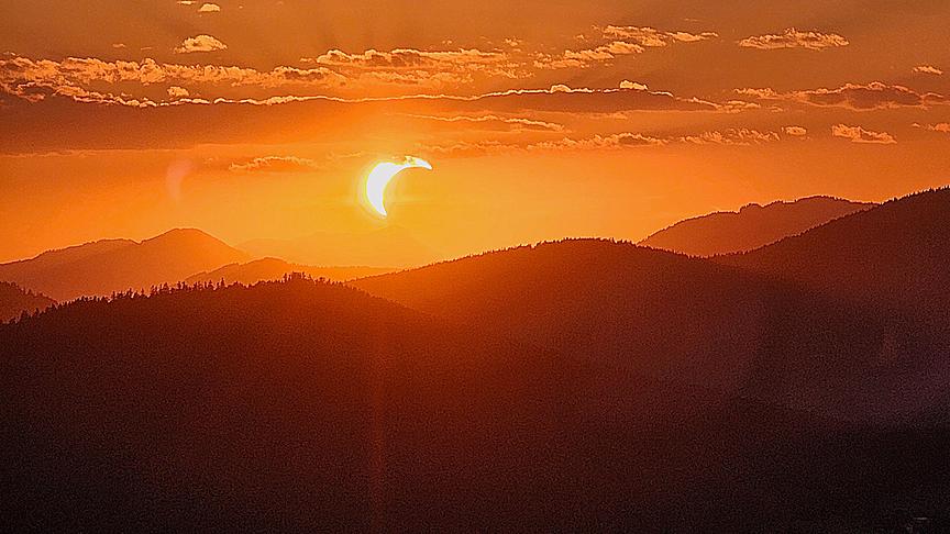 Partielle Sonnenfinsternis in warm orangem Licht mit wenig Wolken über der Sihouette einer hügeligen Waldlandschaft.