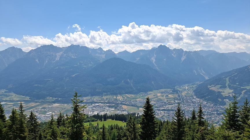 © Erna Linder Ausblick hinab auf ein Tal durch das sich ein Fluss schlängelt und in dem ein Dorf ist. Im Hintergrund sind hohe Berge, über die eine Wolkendecke Richtung Vordergrund zieht.