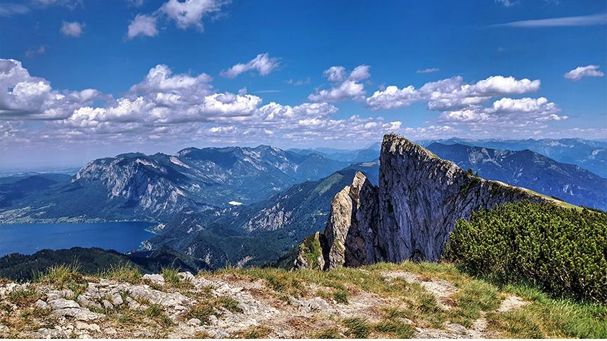 © Emma Sommeregger Aufblick von einem Berg hinab auf einem See und anderere Berge bei Sonnenschein. Der Himmel leuchtet blau und hat einige Schönwetterwolken.