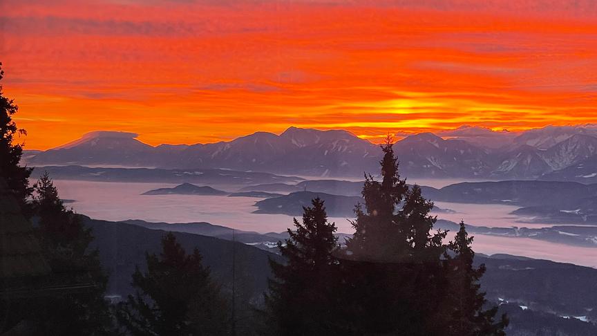 Leuchtend orange-roter Himmel über den Bergen, während im Tal der Nebel hängt.
