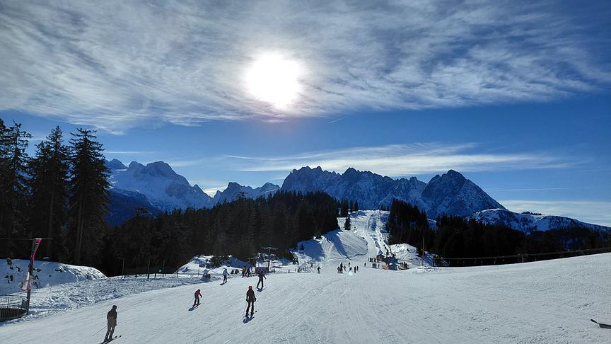 Die Sonne scheint durch Schleierwolken und über den Bergen hinweg auf die gut besuchte Skipiste.
