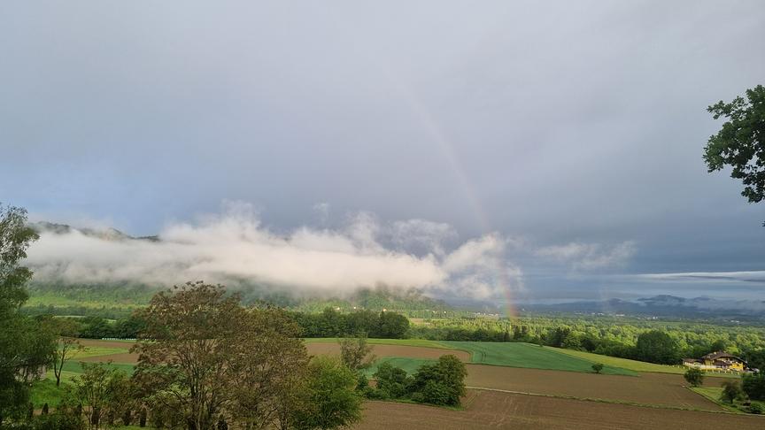 zarter Regenbogen über Felder, Wiesen und Wäldern bei grauer, wolkenverhangener Stimmung.