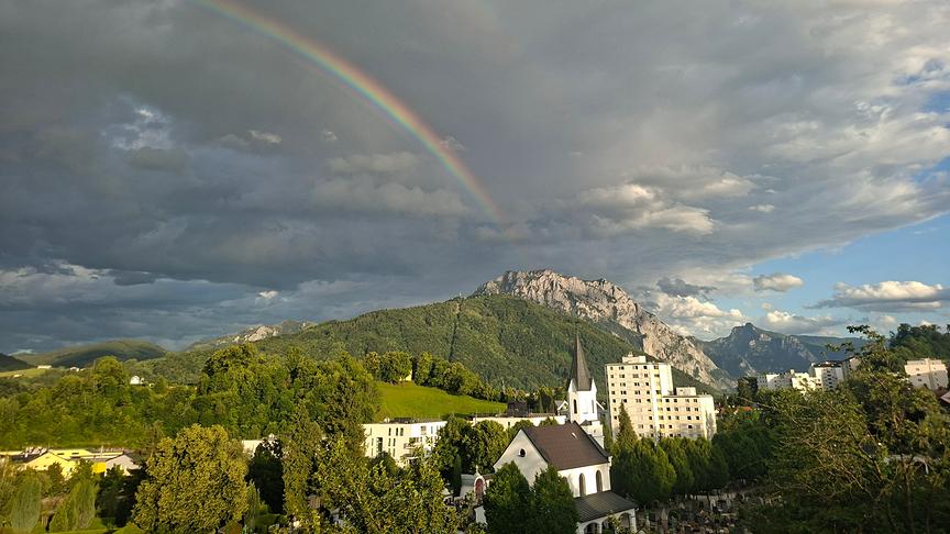 Eine Landschaftsaufnahme bei wechselhaftem Wetter mit einem markanten Regenbogen, der sich vor dunklen Gewitterwolken abzeichnet. Im Hintergrund ragt ein felsiges Bergmassiv empor, dessen Hänge teils bewaldet sind. Im Vordergrund erstreckt sich eine grüne Hügellandschaft mit Bäumen, einer weißen Kirche mit spitzem Turm sowie weiteren Wohngebäuden unter einem teils aufklarenden, blauen Himmel.