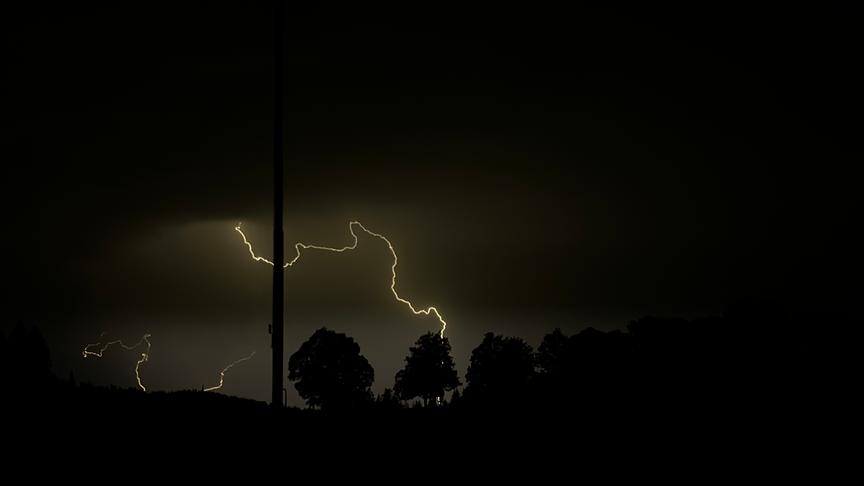 © Gabriele Stöger Dunklere Szenerie in der die Silhouetten von Bäumen und einem Mast zu sehen sind. Ein greller Blitz erhellt die Umgebung, während er gen Boden zischt.