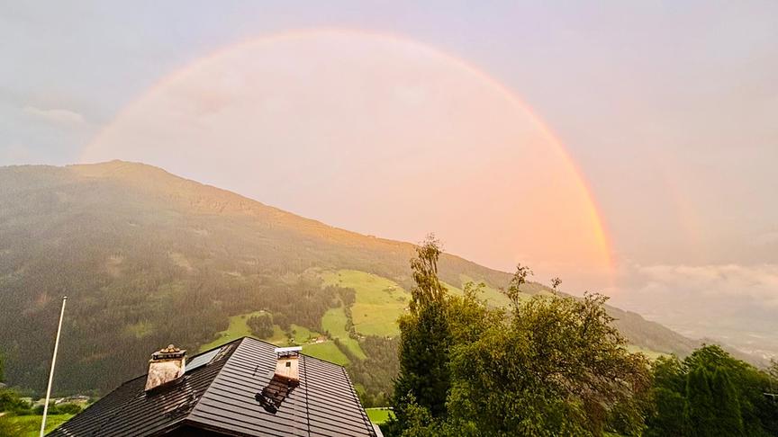 © Isabelle Dorfmann Blick über ein Häuserdach hinweg auf einen grünen Hügel über den sich ein Regenbogen spannt. Unterhalb des Regenbogens scheint warm-oranges Licht, das sich deutlich vom Licht außerhalb des Regenbogens abhebt. Die Atmosphäre ist eher dunstig und die Szenerie daher milchig weichgezeichnet.