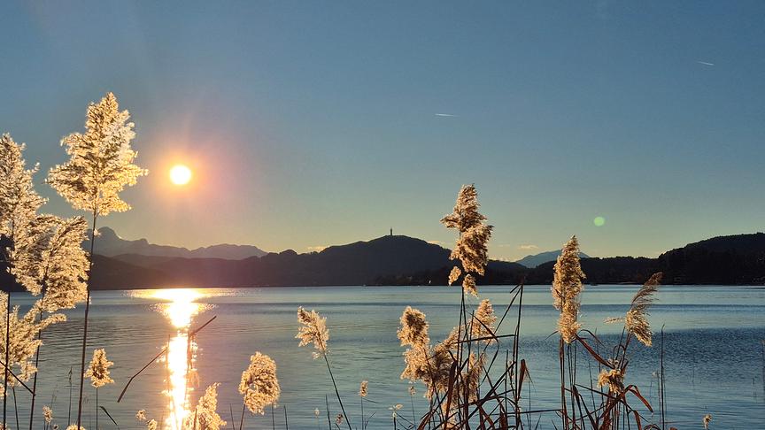 Die Sonne steht tief über den Bergen und dem See, am Ufer wächst Schilf
