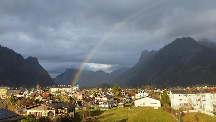 Ein Regenbogen über einer Siedlung, im Hintergrund grau verhangene hohe Berge, im Vordergrund Sonnenschein