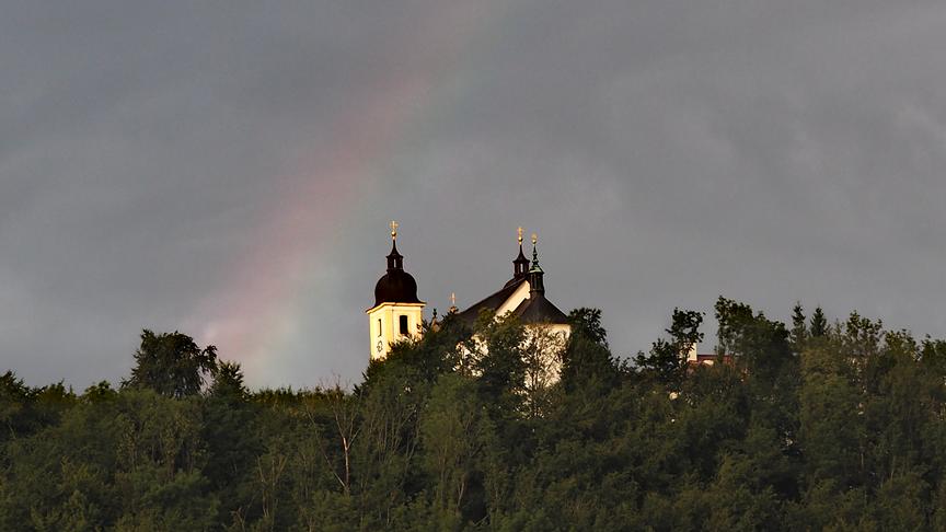 © Christian Behensky Ein schwacher Regenbogen über einer Kirche auf einem bewaldeten Hügel.