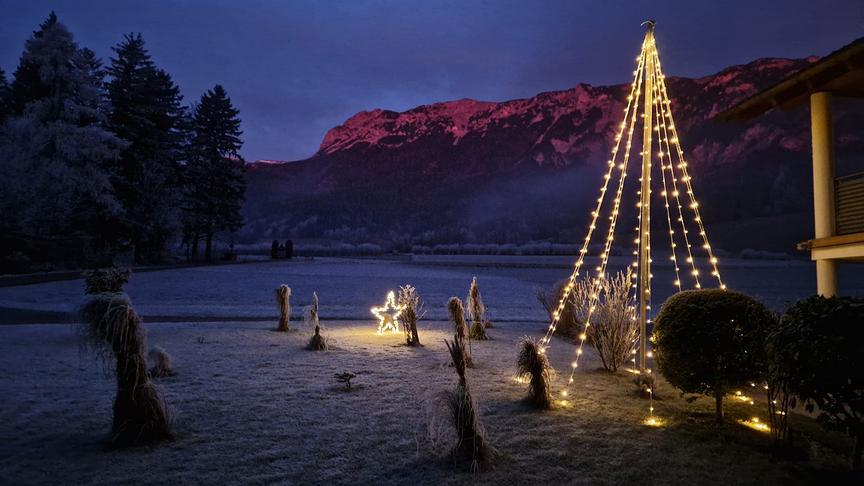 Ein vereister Garten mit Weihnachsbeleuchtung, im Hintergrund Berge, die in der Dämmerung rötlich erleuchtet werden.