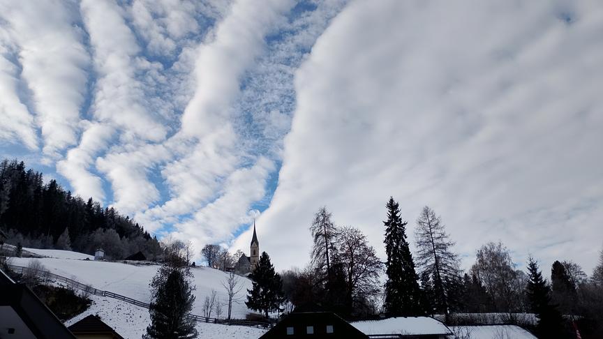 Eine Wolkendecke, die langsam aufbricht und Blick auf blauen Himmel über einer Kirche freigibt.