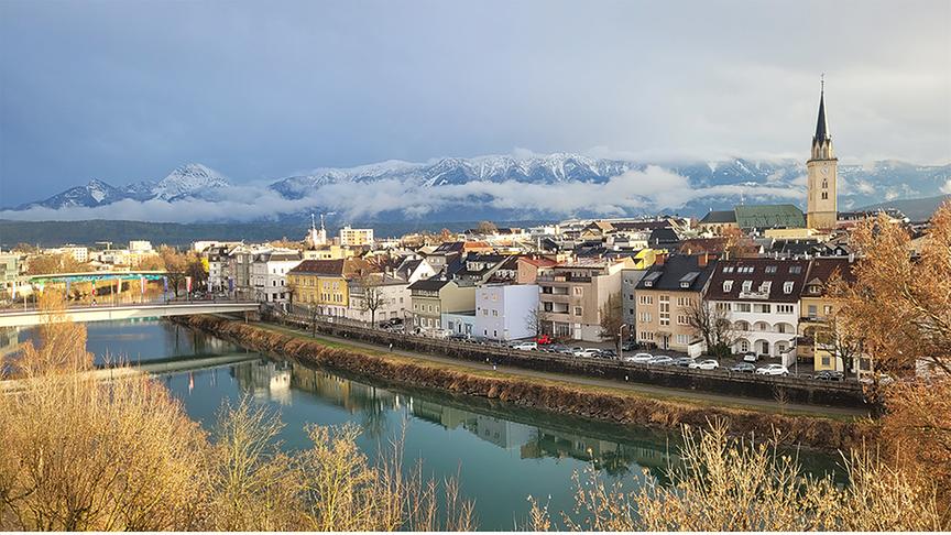 Blick auf einen Fluss und eine Stadt, im Hintergrund wolkenverhangene, verschneite Bergkette