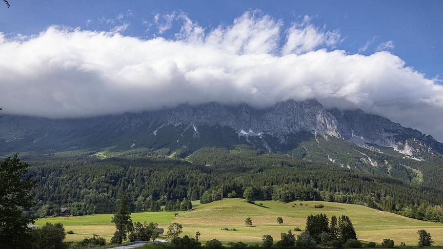 Eine große Wolkendecke stülpt sich über einen Berg. Im Vordergrund Wiesen, Wälder unter blauem Himmel.