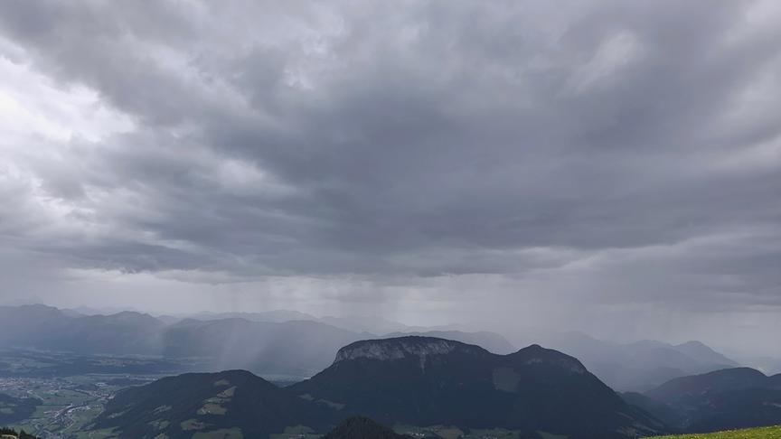 Dunkle Wolken über einer bergigen Landschaft. Regenschleier ziehen sich von den Wolken gen Erde.