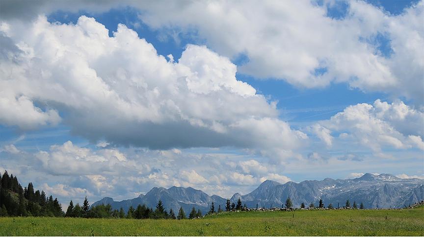 Hohe, bauschige Wolken am blauen Himmel über einem Bergpanorama, Wald und Wiese.