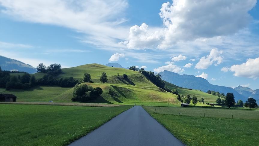 Eine Schönwetterlandschaft mit grünen Wiesen und Hügeln durch die eine geradlinige Straße führt. Im Hintergrund sind einige höher Berge zu sehen.