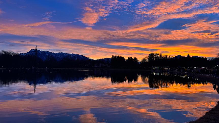 Orangeleuchtender Sonnenuntergang über einem See mit Wald und Bergen im Hintergrund