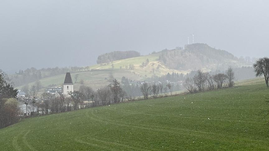 Hügellandschaft mit einer Siedlung, ein Graupelschauer trübt die Sicht und färbt den Himmel grau