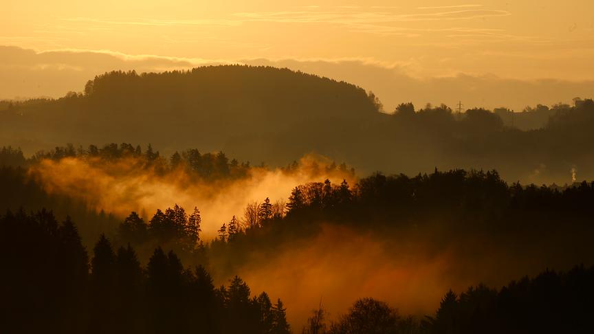 Orange Morgenstimmung mit Silhouetten von bewaldeten Hügeln zwischen denen Nebel hängt