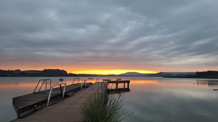 Ein Steg geht weit in einen See hinein. Im Hintergrund sieht man Wälder und Hügel an anderen Ufer. Einige Sonnenstrahlen erhellen einen Streifen dicht über der Erde, der knapp unterhalb der geschlossenen Wolkendecke zu sehen ist und sich zudem im See spiegelt.