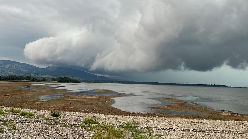 Beeindruckende Wolkenformation breitet sich in Form einer Walze über einem Gewässer, das bereits sehr ausgetrocknet scheint, und Bergen aus.