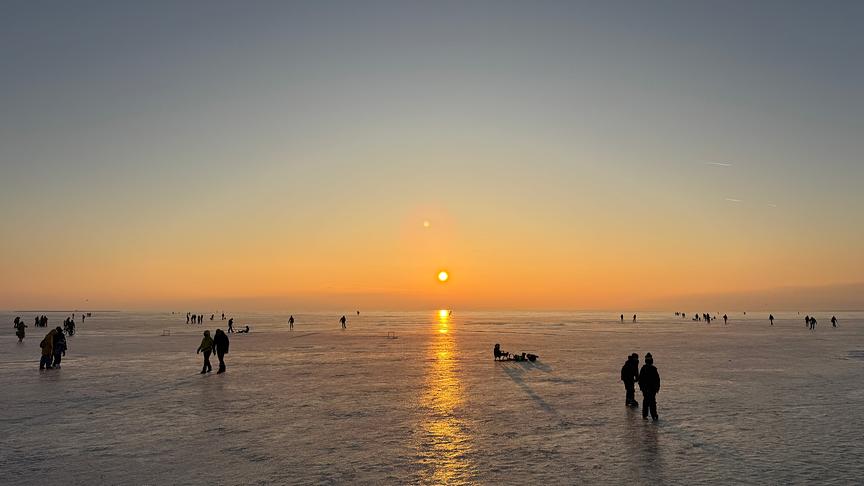 Auf dem zugefrorenen See, der von einer tiefstehenden Sonne in orangen Licht getaucht wird, gehen einige Menschen spazieren.