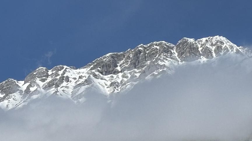 Verschneite Bergkette vor der ein Schreifen Nebel vorbeizieht, am Fuß der Bergkette ist ein Wald