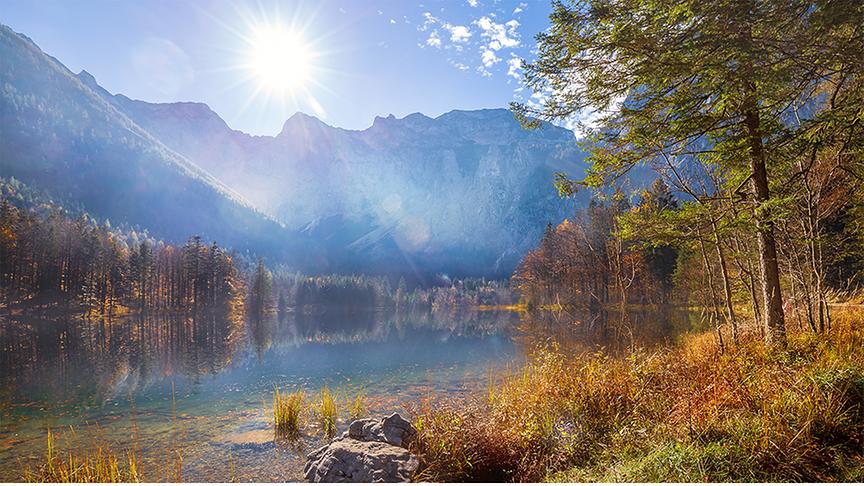 Idyllischer See zwischen hohen Bergen und bereits umgeben von herbstlichen Farben der Bäume am Ufer. Die Sonne scheint hoch über den Bergen im Hintergrund und erleuchtet die Szenerie hell.