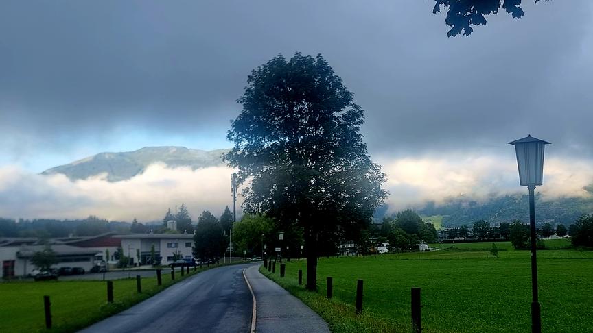 Eine asphaltierte Straße führt links an einer saftig grünen Wiese mit einer traditionellen Straßenlaterne vorbei. Im Hintergrund erstreckt sich eine bergige Landschaft, deren Hänge von einer dichten, tief hängenden Wolken- und Nebelschicht verdeckt werden. Ein großer, laubtragender Baum steht zentral am Straßenrand unter einem wolkenverhangenen, düsteren Himmel.