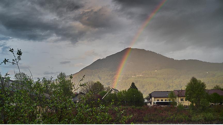 Regenbogen bei dunkler Wolkenstimmung vor einem Berg und über einem Ort.