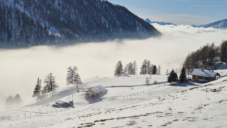 Blick vom Berg auf verschneite Wiesen und Berge sowie dichten, weißen Nebel
