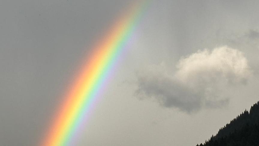 Graue Szenerie mit fast farblosem, leicht bewölktem Himmel und den Silhoetten zweiter bewaldeter Berghänge. Ein Regenbogen spannt sich quer über das Bild und bildet einen farbenprächtigen Kontrast. 