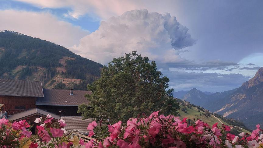 Aufnahme einer sich hoch auftürmenden Wolken zwischen zwei Bergabhängen. Im Vordergrund sind ein Baum, Häuser und vor allem Blumen zu sehen. Die Szenerie scheint von einem Balkon mit Balkonkästen aus gemacht geworden zu sein.