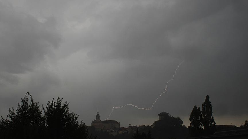 Dunkle Kulisse mit grauen Wolken und dunklen Silhouetten einer Kirche und Bäume. Ein Blitz zieht sich von den Wolken zur Kirche hinunter.