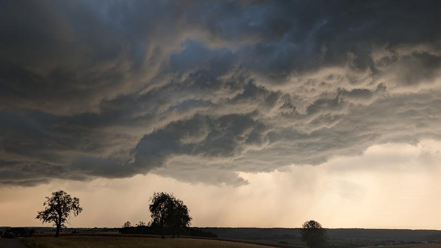 Bedrohlich wirkende, tief hängende Wolkendecke, die sich durch Regen in Schlieren zu Boden zieht. Die darunterliegende Landschaft ist sehr dunkel und nur einige Silhouetten von Bäumen sind zu sehen.