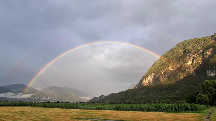 Regenbogen bei bewölktem und grauen Himmel spannt sich über Felder und Wiesen und zwischen Bergen auf.