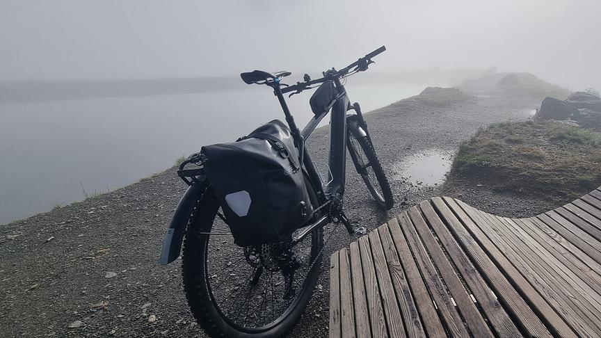 Ein Fahrrad steht neben einer hölzernen Liegebank am Ufer eines Gewässers. Die Atmosphäre ist nebelig und nass.