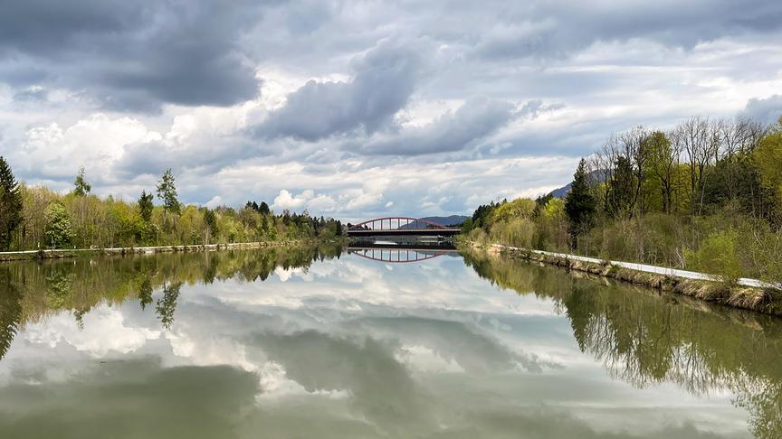 Ein ruhiger Fluss mit bewaldetem Ufer und einer Brücke in der Ferne. Die Wolken am Himmel spiegeln sich im Wasser.