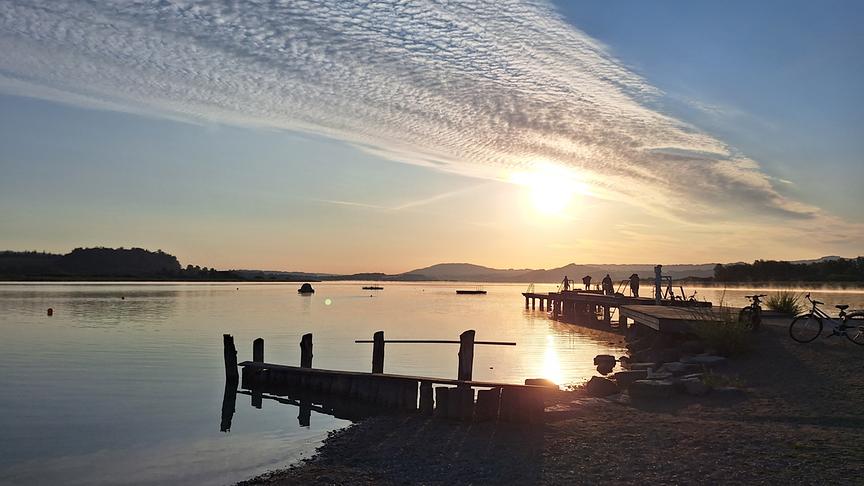 Gewässer in der tiefstehenden, warmen Sonne. Auf einem Steg, der ins Wasser führt stehen einige Menschen. Auf der anderen Seite der Sees ist bewaldetes, leicht hügeliges Ufer. Über den Himmel zieht sich ein Streifen aus vielen kleinen Wolken.