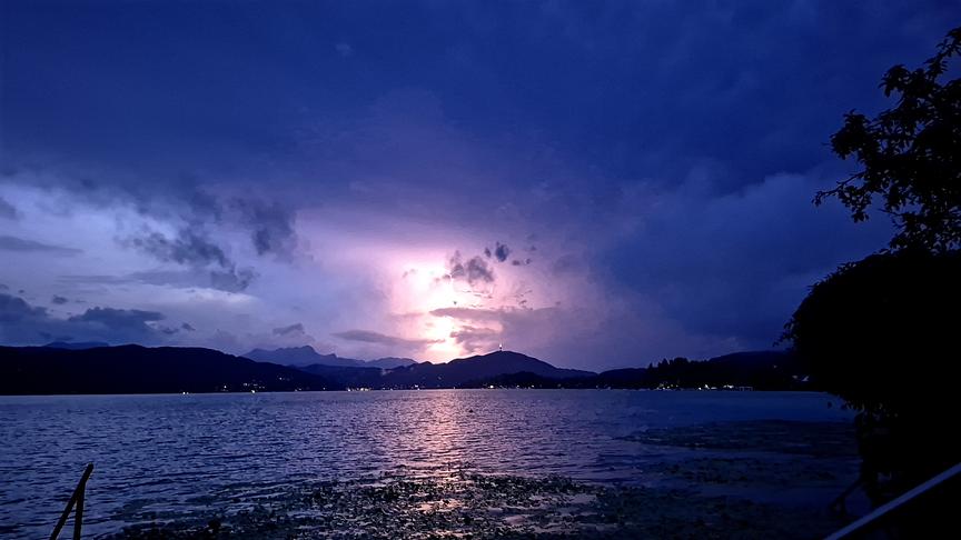 Dunkle Abendstimmung mit Gewitterwolken über einem Gewässer. Im Hintergrund erhellt ein Blitz die Wolken. Die Szenerie spiegelt sich auf der unruhigen Wasseroberfläche.