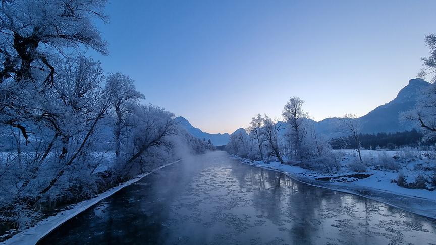 Fluss umgeben von blauer, eisiger Kulisse aus Bäumen und Bergen.